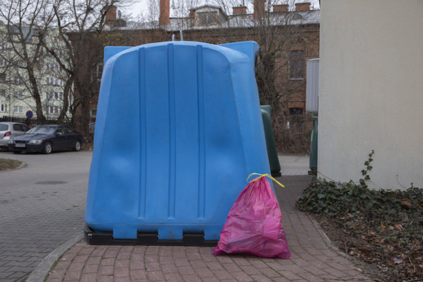 粉色垃圾袋放在城市居民区的一个大蓝色回收箱旁边。塑料垃圾袋放在人行道上，靠近分类回收箱的垃圾处理场景。图片下载