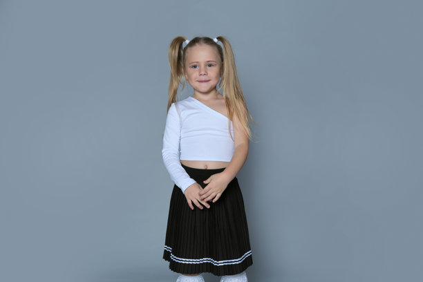 一位金色双马尾、露齿笑的年轻女孩，身穿白色单肩上衣和黑色百褶裙。照片展现了她俏皮无忧的灵魂，捕捉了童年的欢乐瞬间。图片下载