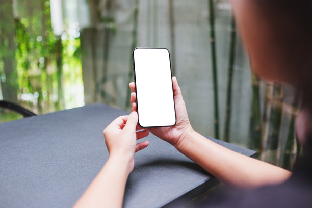 咖啡馆里，女子手持手机并使用，背景为空白桌面屏幕的样机图图片下载