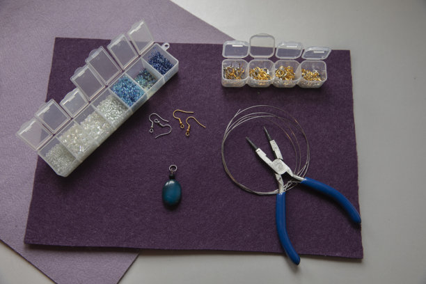 珠宝制作工具，包括钳子、串珠线、金色小搭扣、耳钩和装在塑料盒里的小珠子，摆放在紫色毛毡上，展示了DIY手工爱好。图片下载