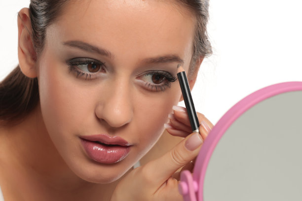 一位深色头发的女性在白色背景下，手持眉笔对着镜子修饰眉毛的棚拍照片图片下载