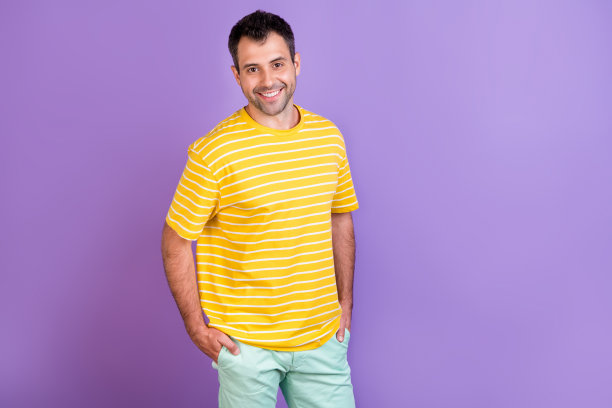 照片：一个年轻男子带着快乐积极的微笑，双手插在口袋里，自信地站在紫色背景前图片下载