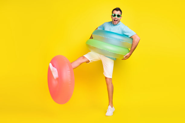 照片：一位穿着蓝色T恤、戴着深色眼镜的英俊帅哥，脸上带着笑容，在黄色背景前玩充气环图片下载