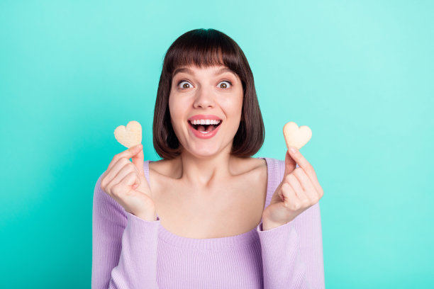 照片肖像，年轻女孩做出两个心形手势，惊讶地盯着，笑着，孤立在明亮的蓝绿色背景上图片下载