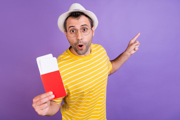 照片：快乐年轻的惊讶男子伸手指着空白处，手持护照，背景为紫色图片下载