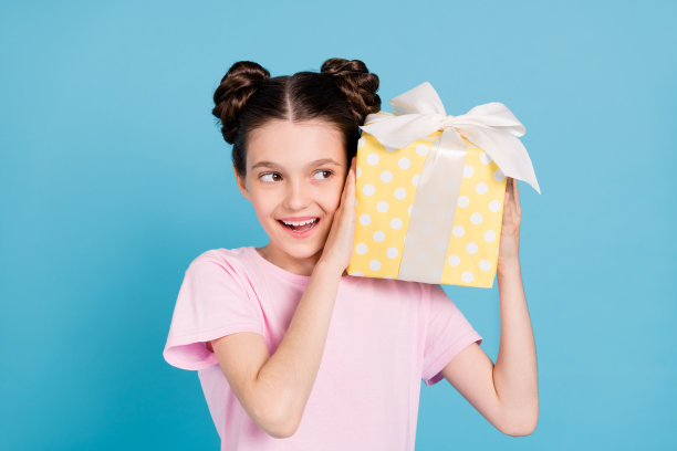 照片：一个梦幻般的小女孩，快乐地望着空白空间，摇晃着一个礼物盒子，好奇地站在蓝色的背景前，孤零零的。图片下载