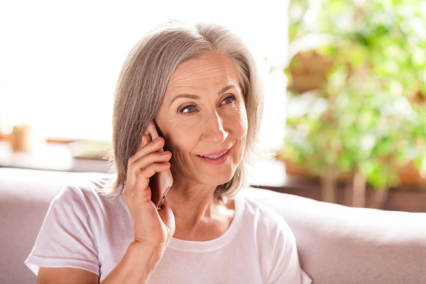 照片：一位漂亮的迷人退休女士，身穿白色T恤，坐在沙发上，微笑着，对着现代小工具说话，在室内房屋的平坦空间里挥手。图片下载