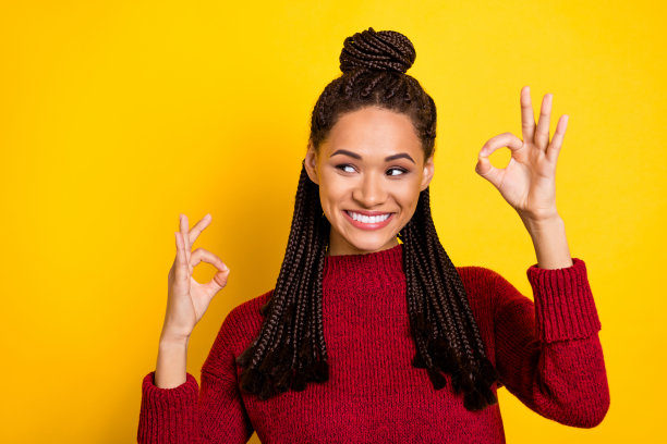 照片：一位漂亮的深肤色女士，穿着红色套头衫，微笑着比出“OK”手势，看向画面外的空白处，背景为黄色。图片下载
