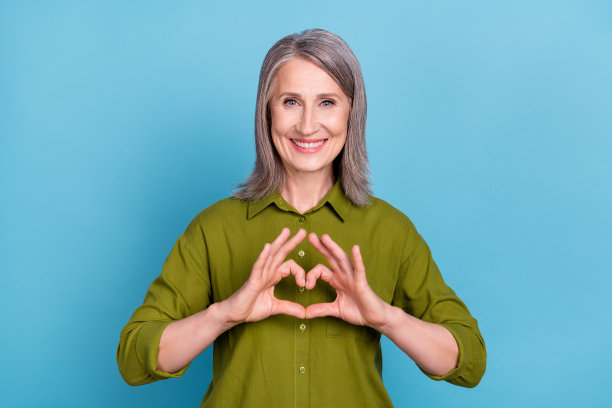 照片：一位浪漫、开朗的退休奶奶，穿着绿色衬衫，在蓝色背景前做出心形手势，竖起大拇指。图片下载