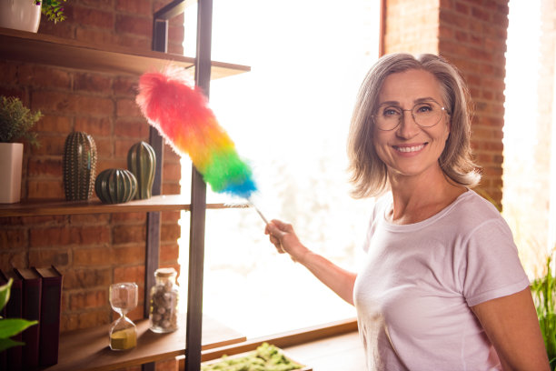 照片：一位穿着白色T恤的可爱、和蔼的老年女性，正在室内打扫架子，脸上带着微笑，场景是她自己的家。图片下载