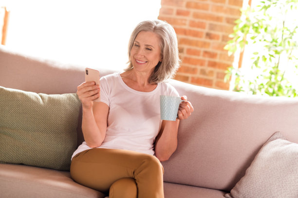 照片：一位穿着白色T恤、可爱迷人的退休女士，坐在沙发上，在家中现代化的室内，一边喝茶一边打字。图片下载