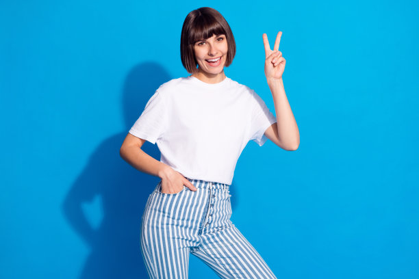 照片肖像，一个留着波波头的漂亮女孩，穿着休闲装，面带微笑，比着V字手势，背景是明亮的蓝色图片下载
