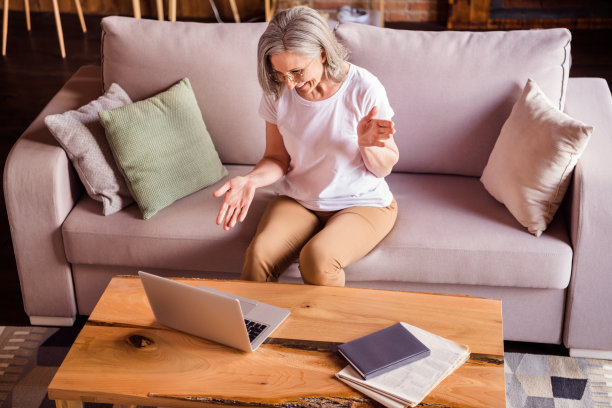 照片：一位穿着白色T恤的甜美迷人的退休女士，坐在沙发上，微笑着使用现代电子设备进行视频通话，室内住宅公寓。图片下载