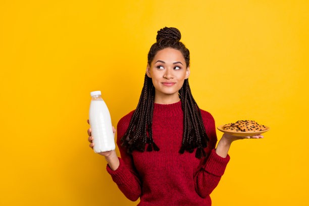 照片：穿着红色毛衣的可爱梦幻黑皮肤女性，手持饼干奶昔，望着虚空，孤立在黄色背景上。图片下载