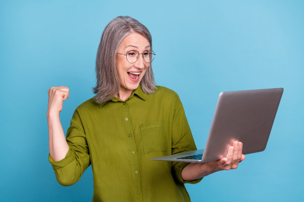 照片：一位戴着眼镜、穿着绿色衬衫的女士，手持上网本，在蓝色背景前惊喜地欢呼图片下载