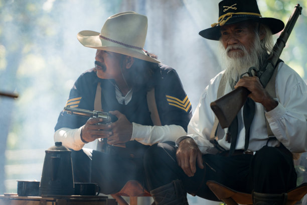 两位牛仔坐在森林营地，手持枪支，烟雾袅袅。图片下载