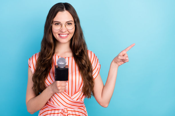 照片：一位戴着眼镜、穿着条纹连衣裙的推广女士手持麦克风，手指着空旷的蓝色背景。图片下载