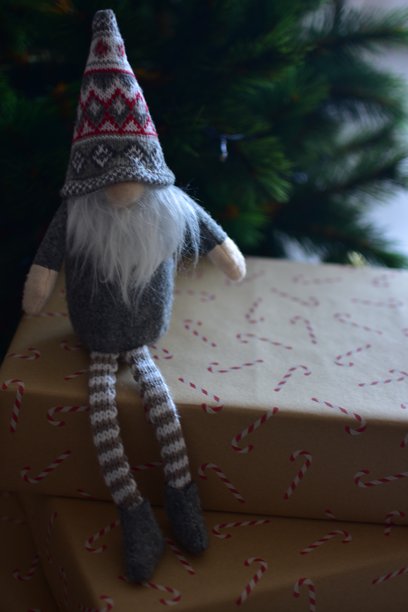 温馨的圣诞场景，一个北欧小精灵坐在一棵装饰过的圣诞树下的礼品盒上。牛皮纸盒、手工制作的软玩具和自然的节日装饰营造出居家温馨的节日氛围。图片下载