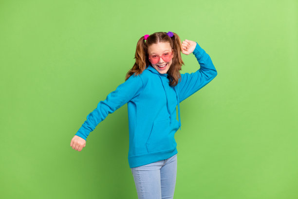 照片：快乐的年轻时尚女孩，心情愉悦地穿着舞服，戴着太阳镜，比心，有趣，孤立在绿色背景上图片下载