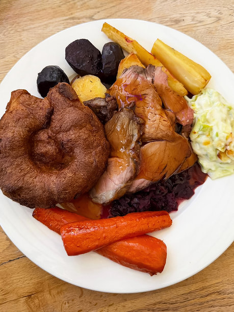 特写图片，展示了周日烤肉晚餐的盘子，包括烤牛肉片、约克郡布丁、芝士韭葱、红卷心菜、烤土豆、胡萝卜、甜菜根、欧防风、肉汁，背景是木桌，俯视角度。图片下载