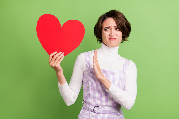 一个女人拿着一张红色心形明信片，做出停止的手势，表情厌恶，背景是柔和的绿色，为照片肖像。图片下载