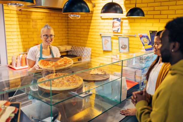 一位穿着围裙的快餐店收银员站在食品柜台后面，两位不同肤色的顾客正在点披萨。这张图片传达了友好的顾客服务和一个典型的快餐环境。图片下载