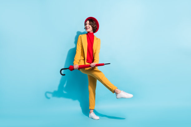 穿着黄色西装外套、红色帽子、可爱俏皮的年轻女士，在蓝色背景前微笑着撑伞跳舞的照片图片下载