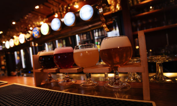 酒吧柜台上摆放着各式各样的精酿啤酒，背景是灯光闪烁的啤酒龙头。图片下载