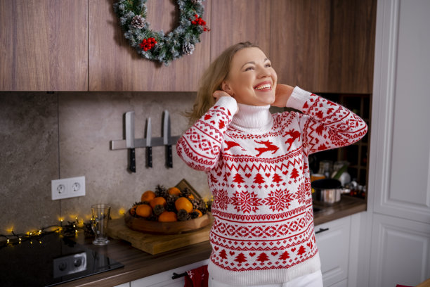 Blond woman in festive red and white christmas sweater leaning on kitchen table in cozy decorated interior with warm lights and greenery, creating joyful spirit, winter mood and cheerful atmosphere图片下载