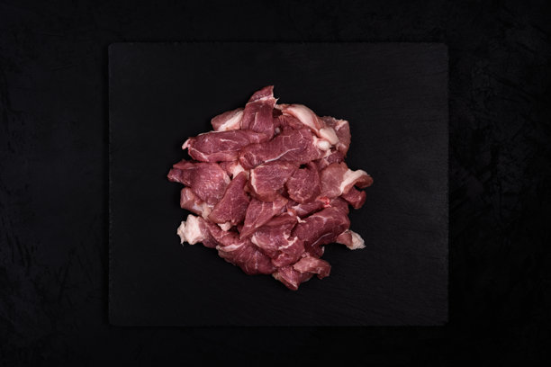 一堆生猪肉炖肉块放在石制砧板上，背景为黑色纹理。新鲜的肉切成块。俯视。中心构图。暗色食物摄影。留白。素材照片图片下载