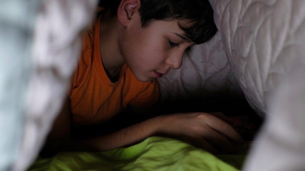 一个穿着橙色T恤的孩子躺在被子里，在昏暗的环境中全神贯注地玩手机。这张照片突显了现代数字习惯和童年的好奇心。图片下载