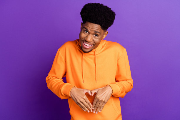 照片：戴着橙色连帽衫的时尚的黑发千禧一代男性，双手比心，孤立在紫色背景上图片下载