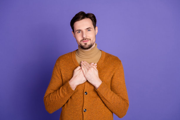 身穿橙色开衫搭配米色高领衫的年轻男子，在紫色背景前摆出心形姿势，适用于生活方式、时尚和广告素材图片下载