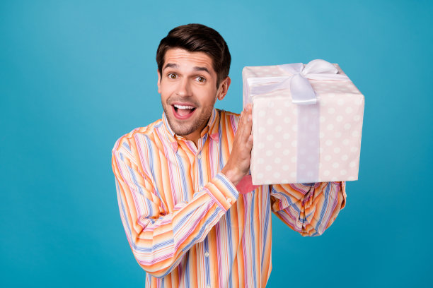 照片：一个年轻兴奋的男人，带着快乐积极的微笑，好奇地拿着一个礼品盒，背景是蓝色的图片下载