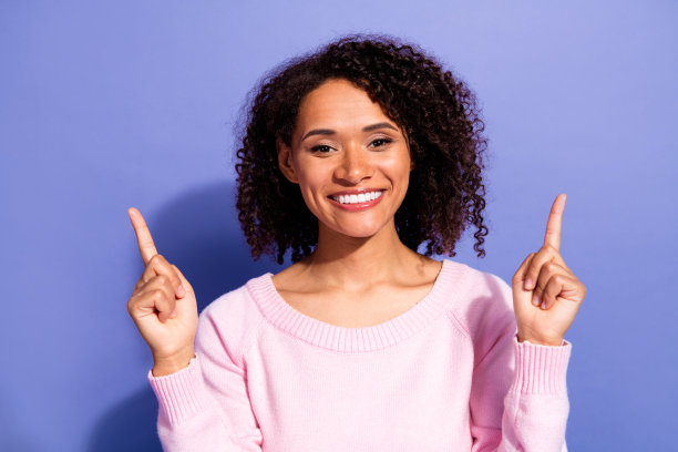 一位年轻女子穿着粉色毛衣，双手向上指着，脸上带着微笑，背景是紫色。这张图片具有休闲生活时尚和积极广告的吸引力。图片下载