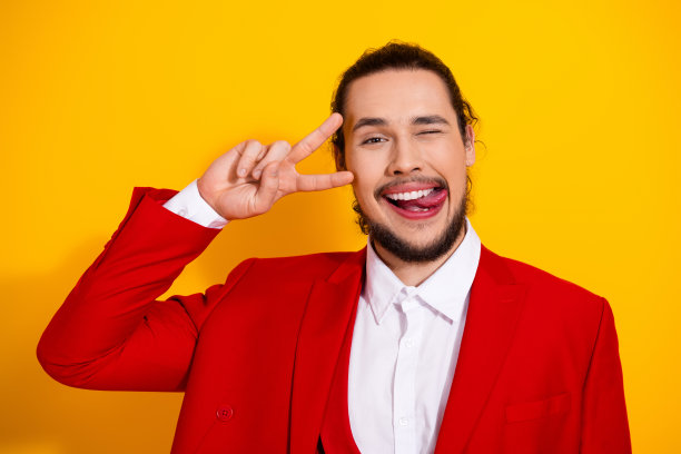 穿着红色西装的时尚滑稽男子，在明亮的黄色背景前摆出胜利手势并吐出舌头，用于时尚素材照片图片下载