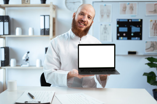 留着胡子的医生穿着白大褂在医院办公室工作，手里拿着一台空白屏幕的笔记本电脑。医疗保健和医学概念。图片下载