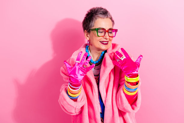 身着粉色仿皮草外套、戴着醒目眼镜的成熟的女性，在粉色影棚内自信微笑，展现魅力、时尚与复古风格，适用于生活方式广告。图片下载