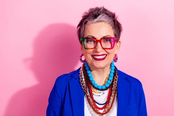 身穿蓝色夹克、佩戴醒目项链和眼镜的自信成熟的女性，背景为粉色，非常适合时尚生活或广告素材。图片下载