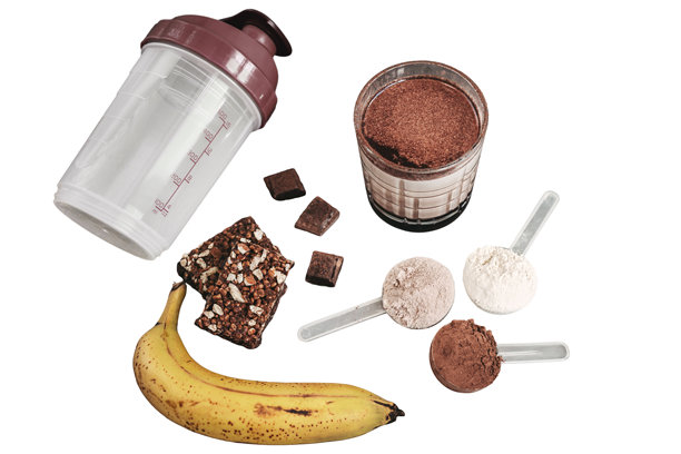 玻璃杯中的蛋白质奶昔，配有三勺乳清蛋白粉、塑料摇摇杯、香蕉、格兰诺拉麦片棒和巧克力碎片，孤立在白色背景上，健身营养概念图片下载