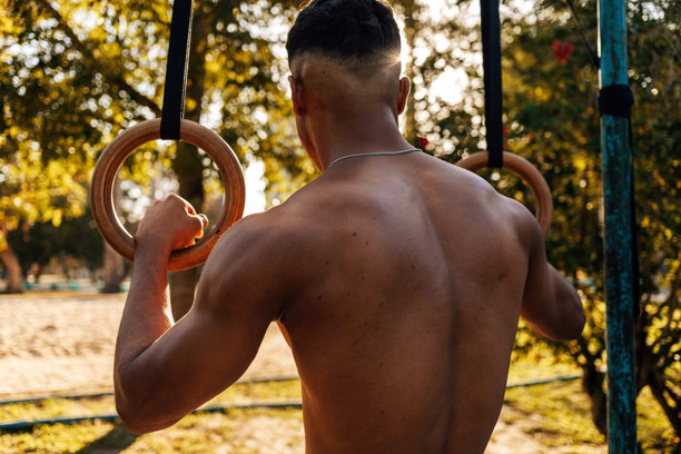 从后面看，肌肉发达的男性运动员在户外抓着体操吊环。这张图片代表了健美训练中暂停或准备的时刻。图片下载