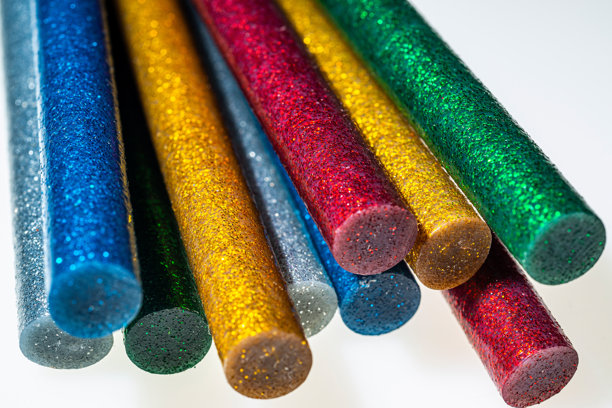 一张宏观照片，展示了涂有闪粉的棍子呈对角线堆叠在白色背景上，突出鲜艳的蓝色、金色、红色、绿色和银色，棍子末端呈圆柱形，具有闪闪发光的质感和光泽表面。图片下载