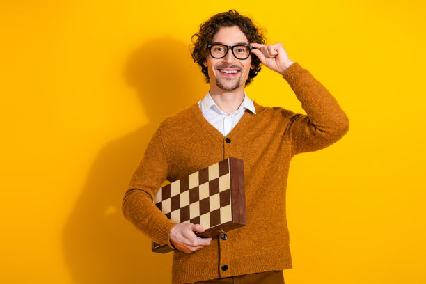 戴眼镜的年轻男子穿着棕色开衫，手持国际象棋棋盘，对着明亮的黄色背景微笑，适用于现代生活方式和广告图片图片下载