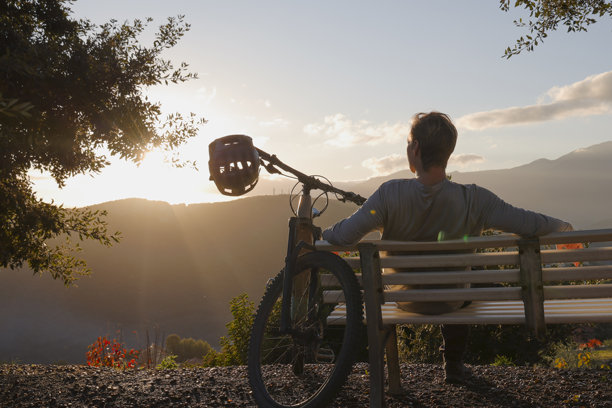 山地车手坐在长凳上，俯瞰山丘，背景是日落图片下载