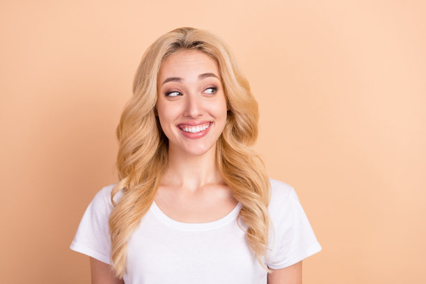 金发女子身穿白色T恤，面带微笑，表情好奇，在柔和的米色背景前，如照片般肖像展示，正在开怀大笑。图片下载