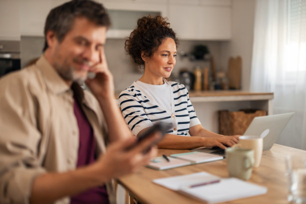 舒适的家庭办公室里，女人在厨房餐桌上用笔记本电脑工作，男人在附近打电话图片下载