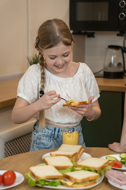 女孩小心翼翼地将黄色芥末酱涂抹在白面包片上，在厨房的料理台上准备三明治，旁边还摆放着蔬菜和火腿等配料。竖幅照片图片下载
