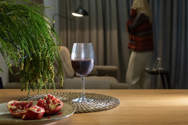 一张玻璃杯红酒、石榴和绿色冷杉枝的照片，放在木桌上，背景虚化，室内有一位正在打电话的女士。图片下载