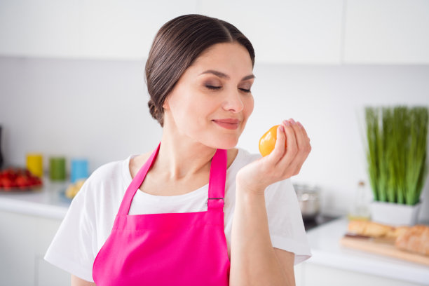 照片：一位留着棕色头发的年轻女子，身穿白色T恤和围裙，在厨房里拿着一个西红柿，脸上露出惊讶的表情。图片下载