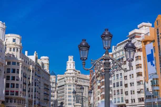 这张图片展示了瓦伦西亚华丽的市政厅广场，通过一盏古董街灯的视角进行拍摄。背景突出了宏伟的新古典主义立面和明亮的蓝色地中海天空。图片下载